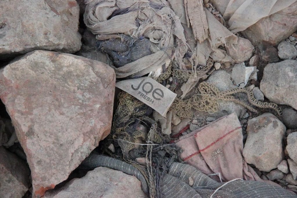 Grey rocks of varying sizes along with parts of grey torn cotton material are mixed with pieces of old wood and string. A dirty Joe Fresh label sits in the middle of the rocks on the ground.